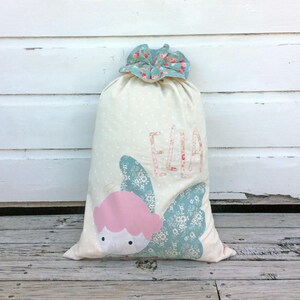 May include: A white fabric gift bag with a blue and pink angel design and the name "Ella" embroidered on the front.