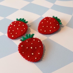 May include: Three felt strawberry pins with green leaves and gold seed details. The strawberries are red and have a white outline.