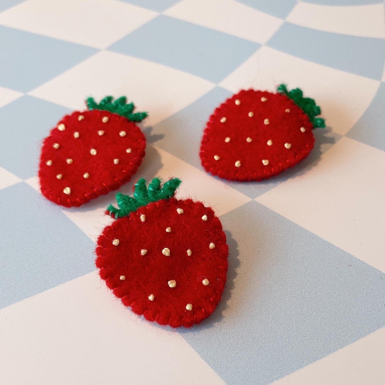 May include: Three felt strawberry pins with green leaves and gold seed details. The strawberries are red and have a white outline.