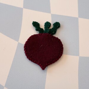 May include: A felt beet with a green felt stem and leaves. The beet is a deep burgundy color and the leaves are a bright green.