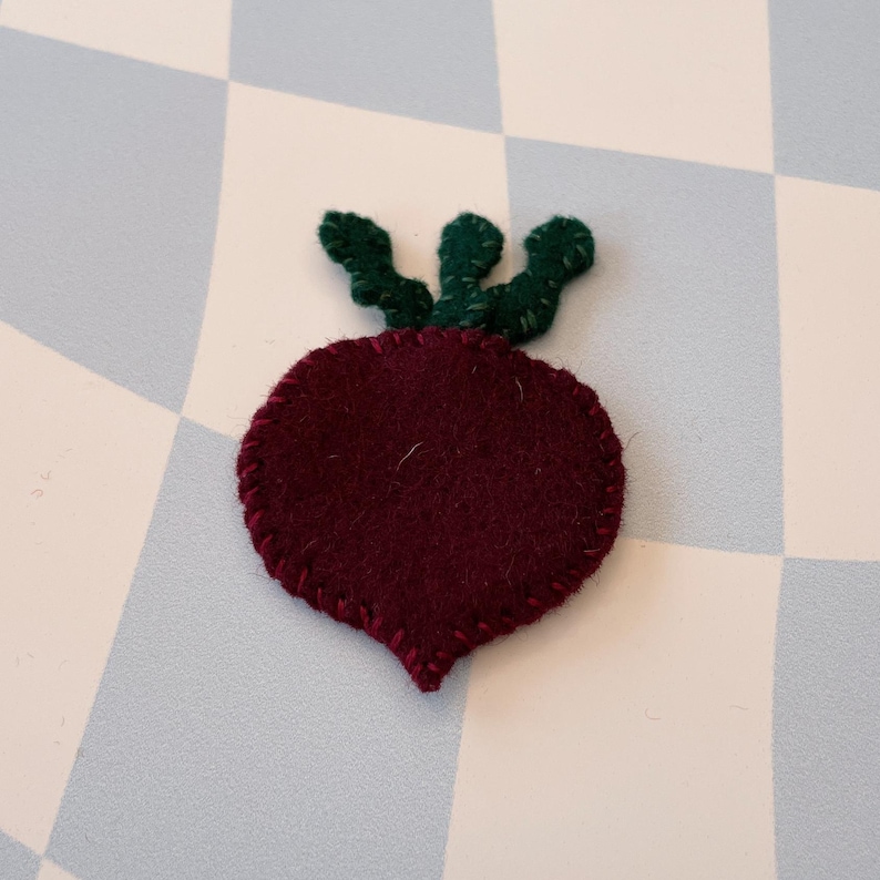 May include: A felt beet with a green felt stem and leaves. The beet is a deep burgundy color and the leaves are a bright green.