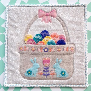 May include: A felt Easter basket with a pink bow, filled with colourful eggs with numbers on them. Two blue felt bunnies are sitting below the basket with flowers and a pink flower in between them.