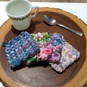 May include: Four colorful woven pot holders on a wooden surface. The pot holders are made of fabric strips woven together in a checkered pattern. The pot holders are in shades of blue, pink, green, and purple.