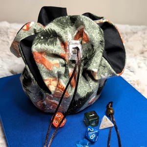 May include: A fabric dice bag with a forest-themed pattern, featuring green, orange, and black accents. A drawstring closure is visible. Several dice in various colors, including blue, green, and orange, are scattered around the bag on a blue surface.