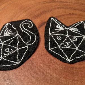 May include: Two black felt cat-shaped patches with white embroidered geometric designs. The cats have a triangular face with eyes, nose, and mouth.