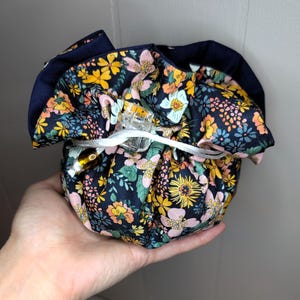 May include: A floral print drawstring pouch in navy blue, yellow, pink, and green. The pouch is held in a hand and secured with a white drawstring and a clear clip. The pouch is designed to hold small items.