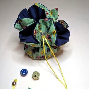 May include: A drawstring dice bag with a teal and navy blue floral pattern. The bag is open, revealing a navy blue interior. Four multi-sided dice and a yellow drawstring are visible. The bag is designed for tabletop gaming.
