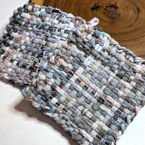 May include: Two woven potholders in shades of gray, blue, and pink. The potholders are rectangular with a woven texture and a loop for hanging. The potholders are handmade and appear to be made of fabric scraps.