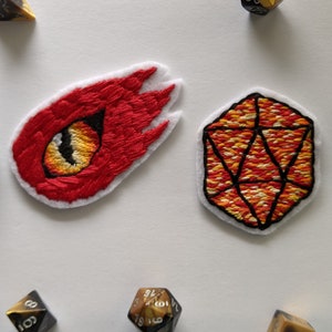 May include: Two embroidered patches, one depicting a red and yellow fire with an eye, and the other a red and yellow dice with black outlines.