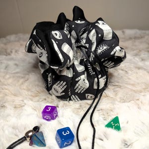 May include: Black and white drawstring dice bag with a repeating pattern of hands and eyes. Several multi-sided dice in purple, blue, and green are scattered around the bag. A black drawstring is visible.