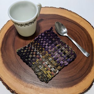 May include: A woven trivet with purple, brown, and yellow yarn on a wooden surface with a white mug and a silver spoon.