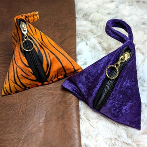 May include: Two triangular pouches. One is orange with black stripes, resembling a tiger pattern. The other is a deep purple. Both have black zippers with gold-colored pulls and a small fabric loop.