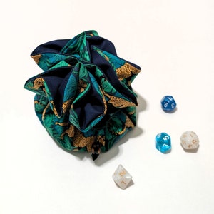 May include: A blue and green fabric dice bag with a floral pattern. The bag is filled with dice, including a blue d6 with the number 4, a blue d6 with the number 7, a white d6 with the number 1, a white d6 with the number 8, and a white d6 with the number 5.