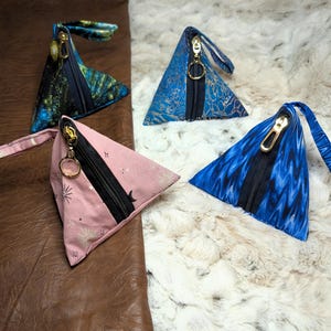 May include: Four triangular zipper pouches in various patterns and colors. One is pink with a star pattern, another is blue with a swirling design, and two others have a blue and green pattern. Each pouch has a black zipper and gold-colored hardware.