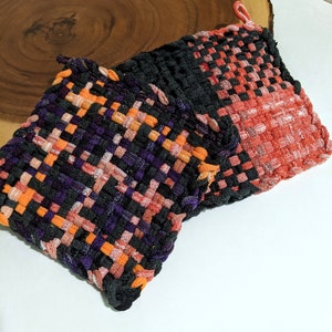 May include: Two woven pot holders in black, orange, and purple. The pot holders are made of a thick, textured fabric and have a checkered pattern.