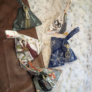 May include: Four triangular fabric pouches with zippers and wrist straps. The pouches feature various patterns, including a gray dinosaur print, a white owl print, a floral design, and a blue snowflake pattern. The zippers are silver and gold.