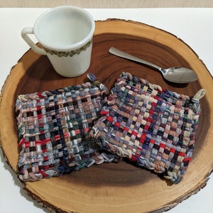 May include: Two colorful woven pot holders on a wooden surface with a white mug and a silver spoon.