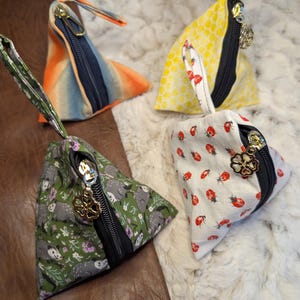 May include: Four triangular fabric pouches with zippers and charms. One is orange and blue, one is yellow with white polka dots, one is green with a floral and animal print, and one is white with red ladybugs. Each has a zipper and a charm.