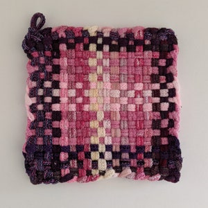 May include: A square, woven hot pad in shades of pink, purple, and cream. The handmade pad features a loop for hanging and a tightly woven pattern. The color gradient transitions from dark purple to light pink.