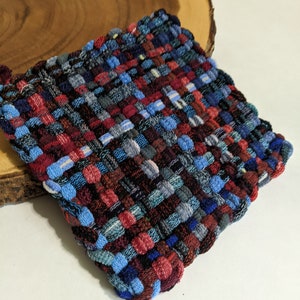 May include: A woven square coaster in shades of blue, red, and black. The coaster is made of a thick, textured fabric and has a slightly frayed edge.