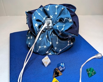 May include: A blue drawstring dice bag with a constellation pattern, white cord, and several multi-sided dice. The dice are various colors, including yellow, white, blue, brown, and green. The bag is on a blue surface.