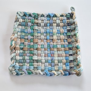 May include: A woven pot holder with a square shape and a textured surface. The pot holder is made of various shades of blue, green, and brown yarn.