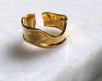May include: A gold-colored adjustable ring with a textured, hammered design. The ring has an open design and sits on a white marble surface. The ring is a piece of jewelry.