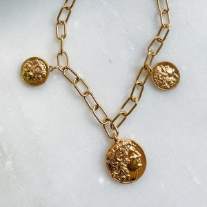 Könnte beinhalten: Eine goldene Kettenkette mit drei goldenen Münzenanhängern, die Profilbilder von historischen Persönlichkeiten zeigen. Die Kette besteht aus ovalen Gliedern und hat einen Perlenschluss.