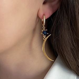 Peut inclure: Boucles d'oreilles créoles dorées avec un motif céleste. Chaque boucle d'oreille présente une petite étoile en pierre précieuse bleu foncé et un pendentif en forme de croissant de lune. Les créoles sont ornées de petits cristaux transparents.
