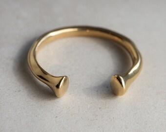 May include: A gold-colored open cuff bracelet. The bracelet has a smooth, rounded band that curves around to two flat, rounded ends. The bracelet is set against a neutral background, highlighting its simple, elegant design. Jewelry.