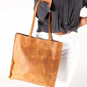May include: A large, brown leather tote bag with a wide, flat base and two long straps. The bag is made of a distressed leather with a natural, rustic look.