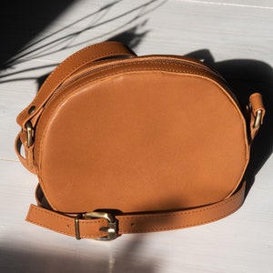 May include: A brown leather crossbody bag with a round shape and a gold buckle. The bag has a long strap and a zipper closure.