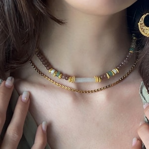 Puede incluir: Un collar en capas con una cadena dorada y un choker de cuentas. El choker tiene cuentas marrones, verdes y blancas con detalles dorados. Una piedra rectangular de color claro está en el centro.