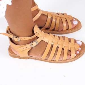 May include: Tan leather gladiator sandals with multiple straps and buckles. The sandals have a flat sole and are made of a soft, supple leather.