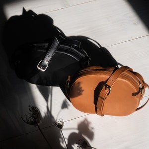 May include: Two leather handbags, one black and one brown, on a white wooden surface. The brown bag is oval shaped and has a strap.