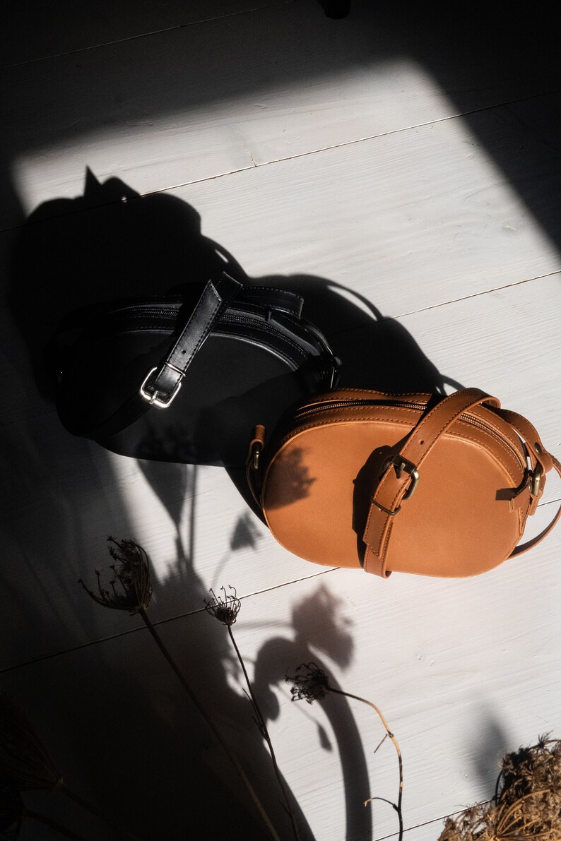 May include: Two leather handbags, one black and one brown, on a white wooden surface. The brown bag is oval shaped and has a strap.