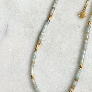 May include: A delicate gold chain necklace with a heart charm and a strand of light blue, white, and brown beads.