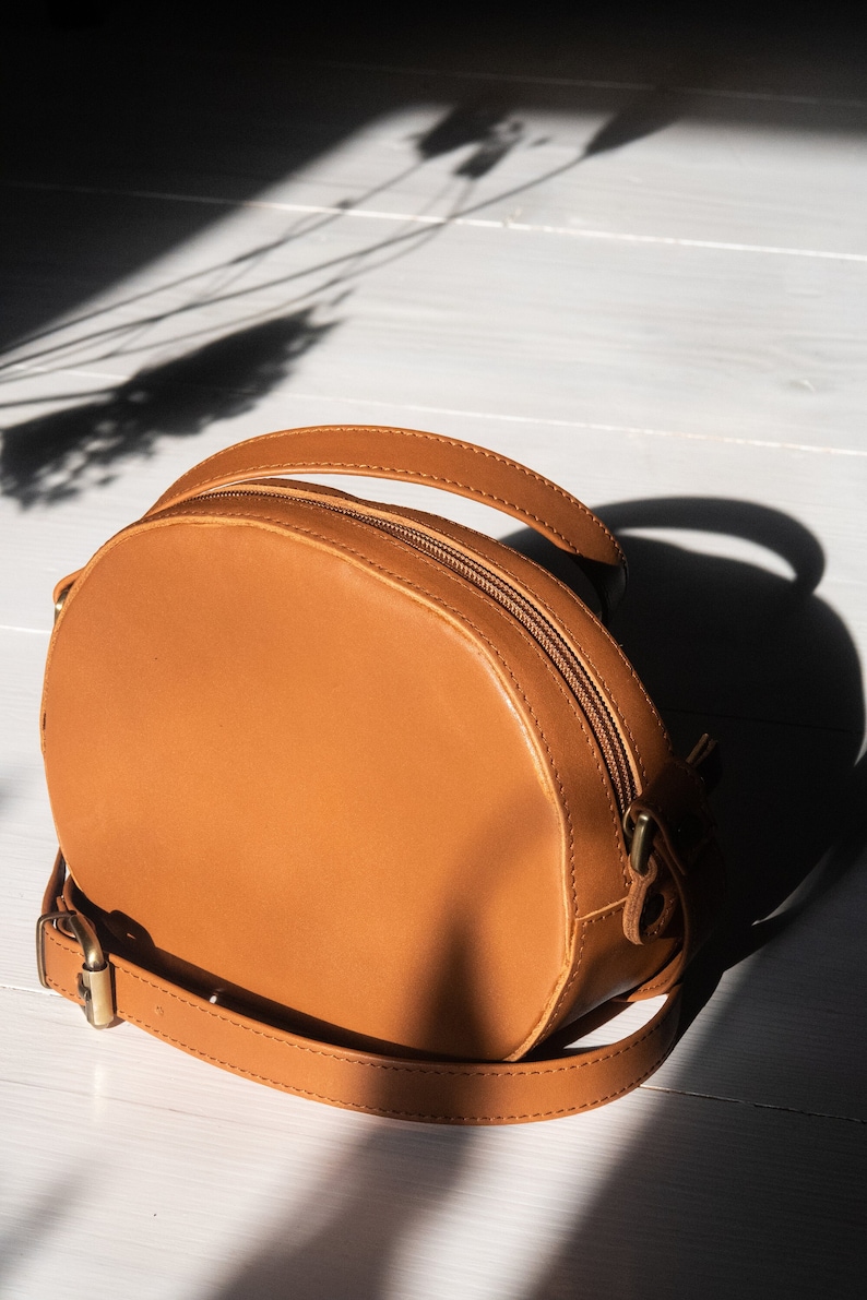 May include: A tan leather round crossbody bag with a zipper closure and a long adjustable strap.