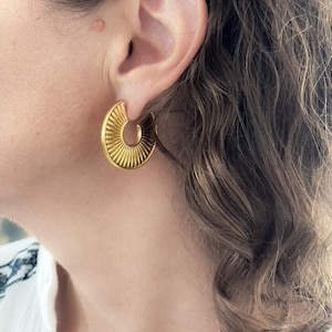 Könnte beinhalten: Goldene Creolen mit Sonnenstrahlen-Design. Die Ohrringe werden von einer Person mit braunen Haaren getragen.