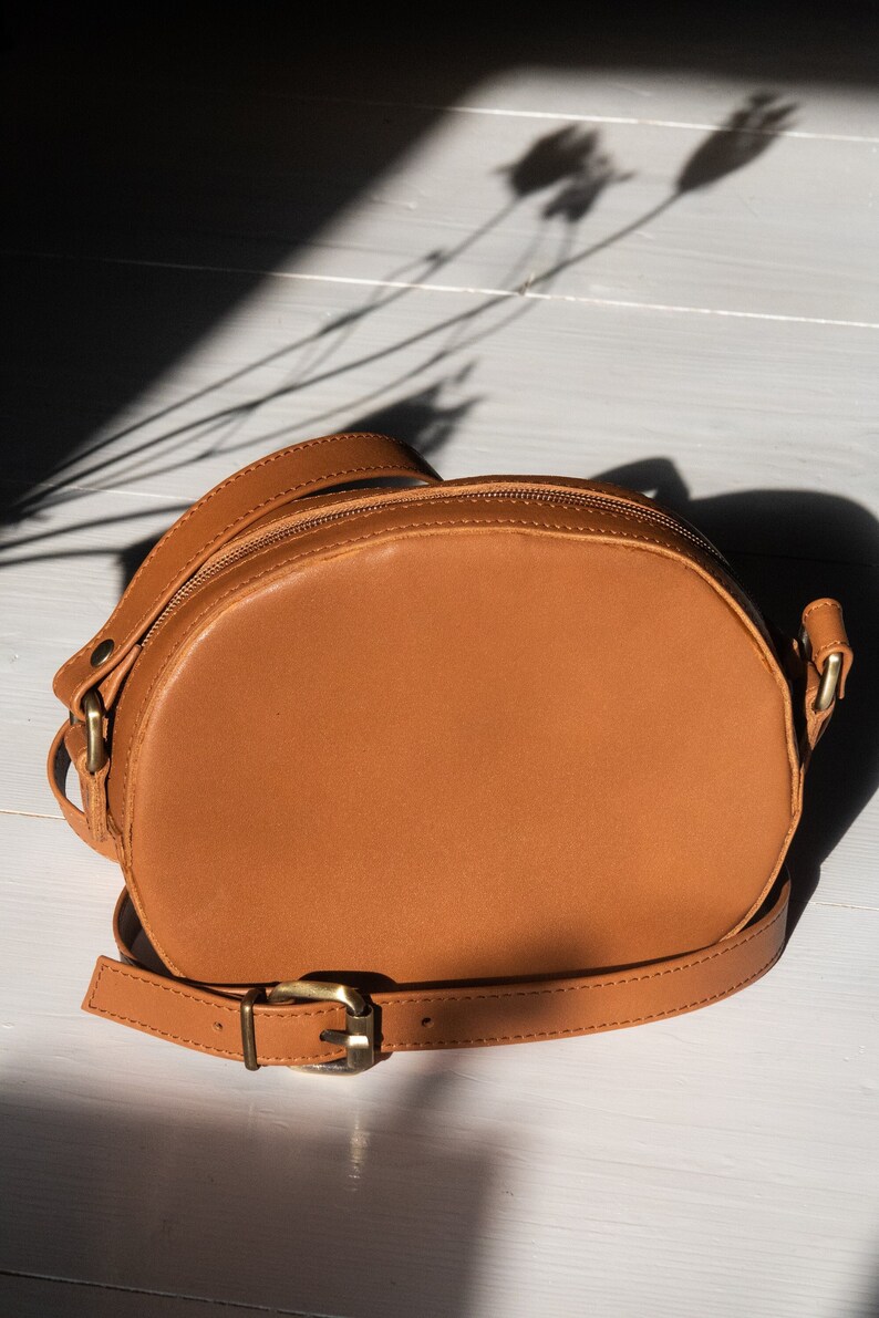 May include: A brown leather crossbody bag with a round shape and a gold buckle. The bag has a long strap and a zipper closure.
