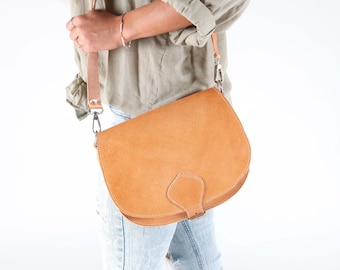 May include: A light brown leather satchel bag with a long adjustable strap. The bag has a flap closure with a buckle and a small, pointed flap on the front.