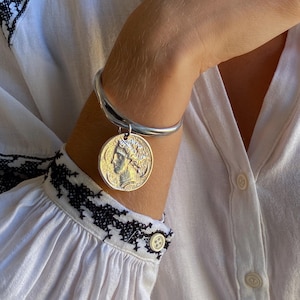 Könnte beinhalten: Ein silberfarbenes Armband mit einem kreisförmigen Band und einem Münzanhänger. Die Münze zeigt das Profil einer Person. Das Armband wird am Arm getragen. Die Person trägt ein weißes Hemd mit schwarzer Stickerei.