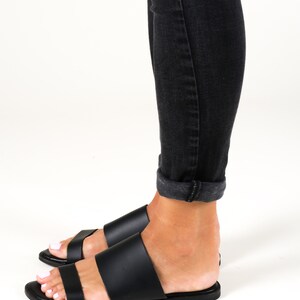 May include: Black leather slide sandals with two straps. The sandals are worn on a person's feet with black jeans.