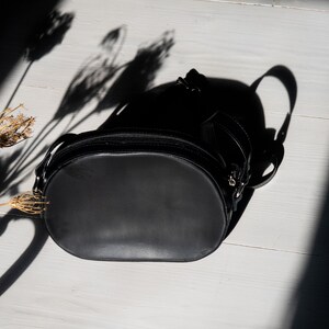 May include: A black leather oval-shaped handbag with a zipper closure and a strap. The bag is on a white wooden surface.