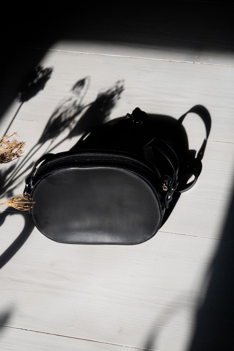 May include: A black leather oval-shaped handbag with a zipper closure and a strap. The bag is on a white wooden surface.