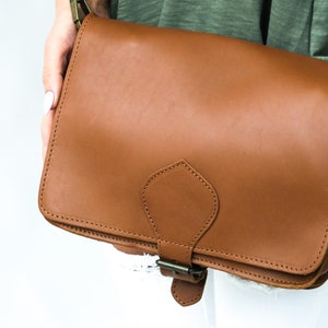 Puede incluir: Un bolso de cuero marr&oacute;n con cierre de solapa y hebilla decorativa. El bolso tiene un dise&ntilde;o cosido y una peque&ntilde;a solapa en la parte delantera.
