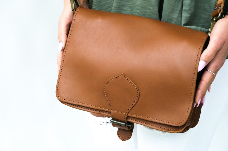 Puede incluir: Un bolso de cuero marr&oacute;n con cierre de solapa y hebilla decorativa. El bolso tiene un dise&ntilde;o cosido y una peque&ntilde;a solapa en la parte delantera.