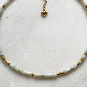 May include: A delicate necklace made with small, light blue, green, and beige beads. The necklace has a gold chain with a small, gold, diamond-shaped pendant.
