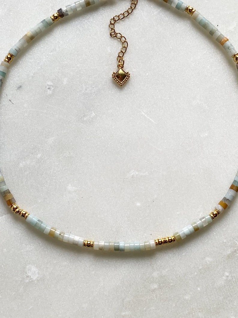 May include: A delicate necklace made with small, light blue, green, and beige beads. The necklace has a gold chain with a small, gold, diamond-shaped pendant.