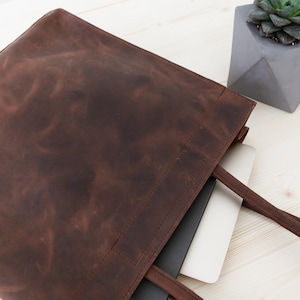May include: A brown leather tote bag with a laptop computer inside. The bag has two long straps and a zipper closure.